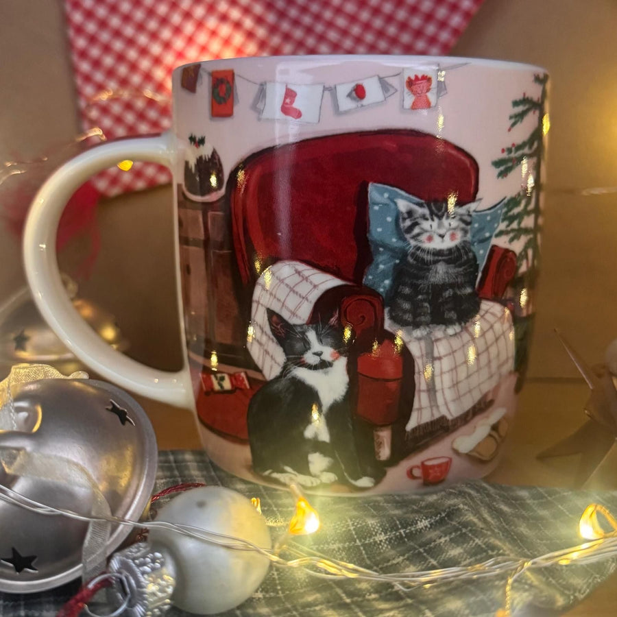 A white ceramic mug decorated with a cosy illustration of two cats lounging in a red armchair under warm fairy lights
