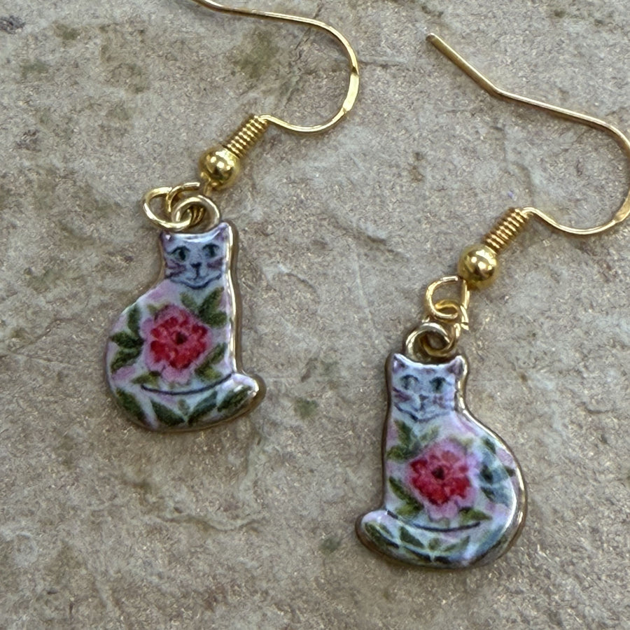 A pair of dangling earrings shaped like seated cats, decorated with pink roses and green leaves on a white background, hanging from gold-tone fishhook wires