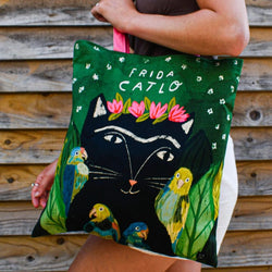 A person holds a large green tote bag featuring a black cat face with pink flowers on its forehead, colorful parrots, and the words 'FRIDA CATLO' in white.