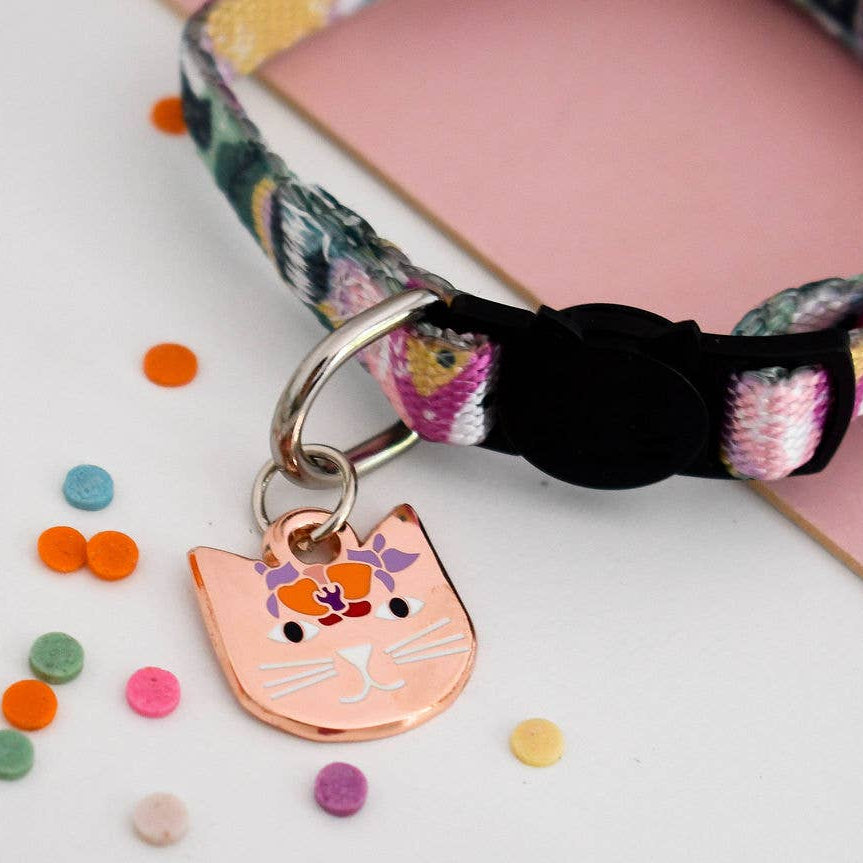 A close-up of a cat collar with a colourful floral pattern, a black buckle, and a peach-coloured cat-shaped metal tag