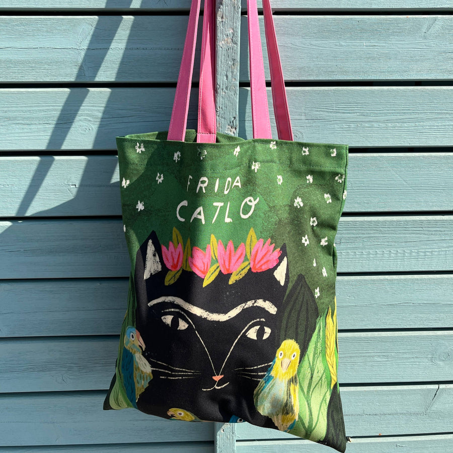 A tote bag with pink handles. The green bag features white text reading 'FRIDA CATLO', a large black cat face, pink flowers, and bird illustrations