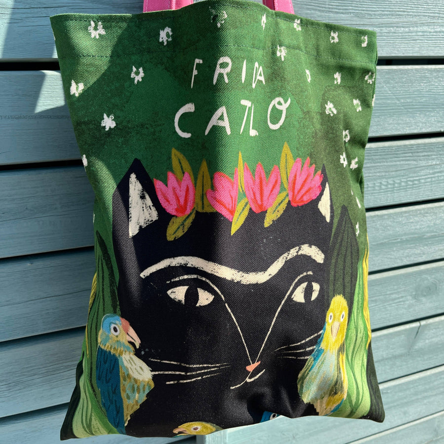 A green canvas tote bag with pink handles, featuring a stylized black cat face, pink flowers, birds, and the text 'FRIDA CATLO'
