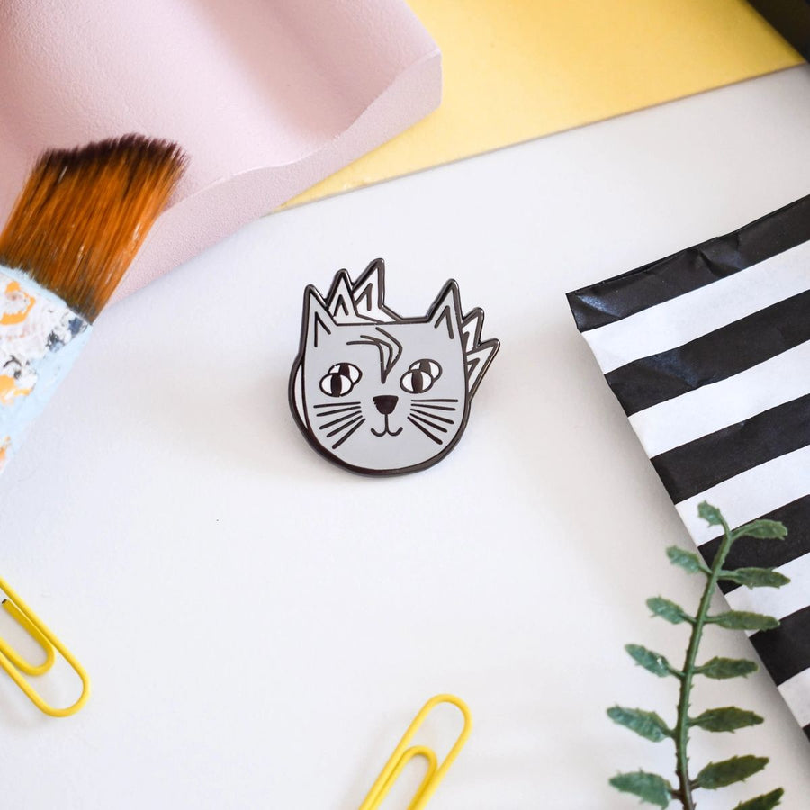 An enamel pin badge with an illustration of a whimsical cat's head