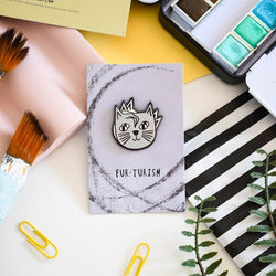 An enamel pin badge with an illustration of a cat's head, placed on a card with 'FUR-TURISM' text