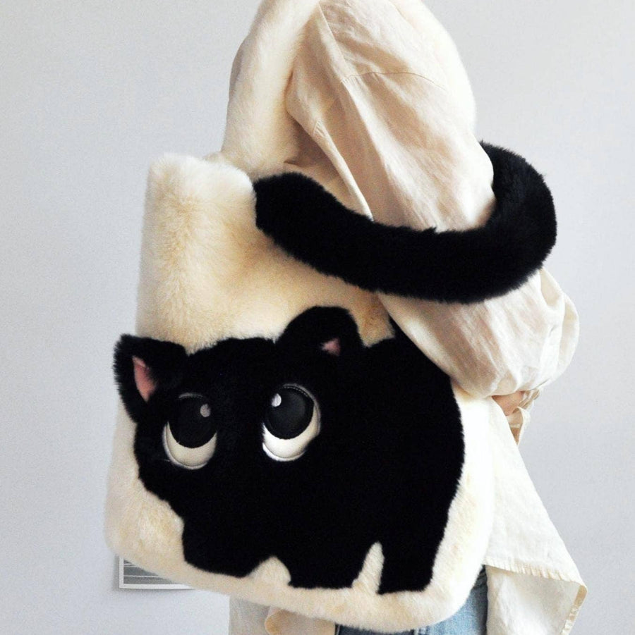 A person carries a plush black-and-cream cat-shaped shoulder bag with large embroidered eyes and a black tail