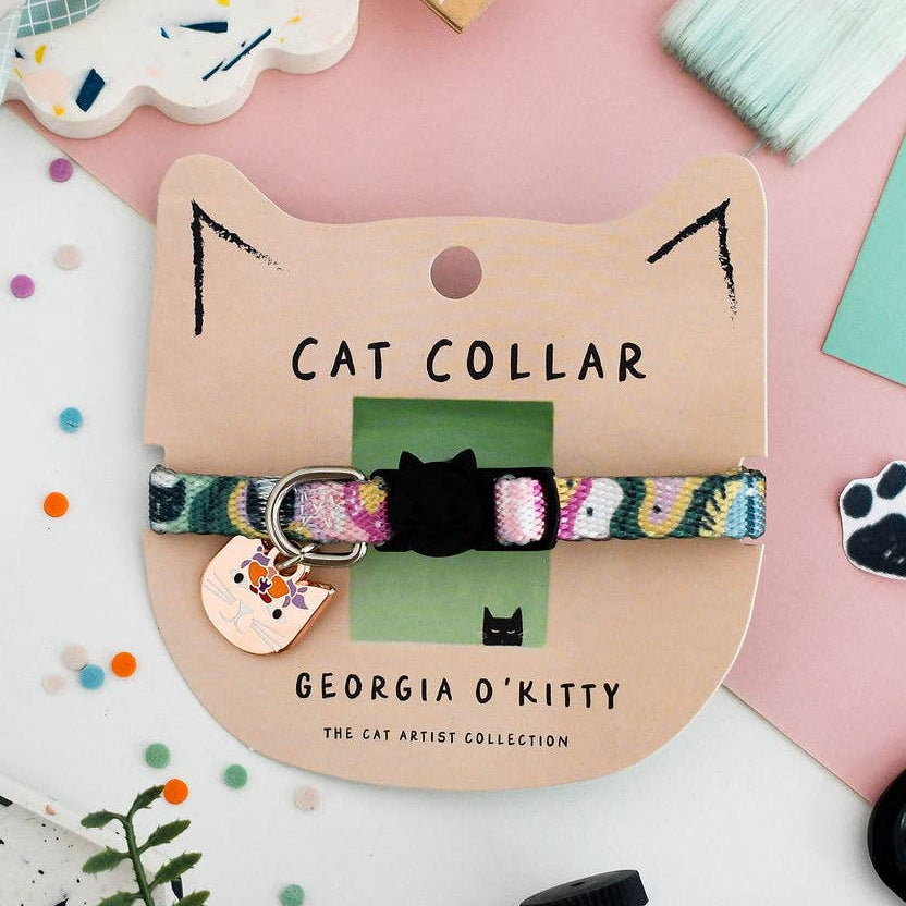 A cat collar displayed on a cardboard cat-shaped card. The collar has a multicolour patterned fabric strap with a metal cat charm