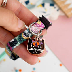 A close-up of a hand holding a colourful cat collar with a cat-shaped metal charm decorated with orange and purple flowers