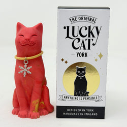 A red Lucky Cat figurine wearing a gold collar with a silver snowflake charm, displayed next to its white and black retail box featuring 'The Original Lucky Cat York' branding.
