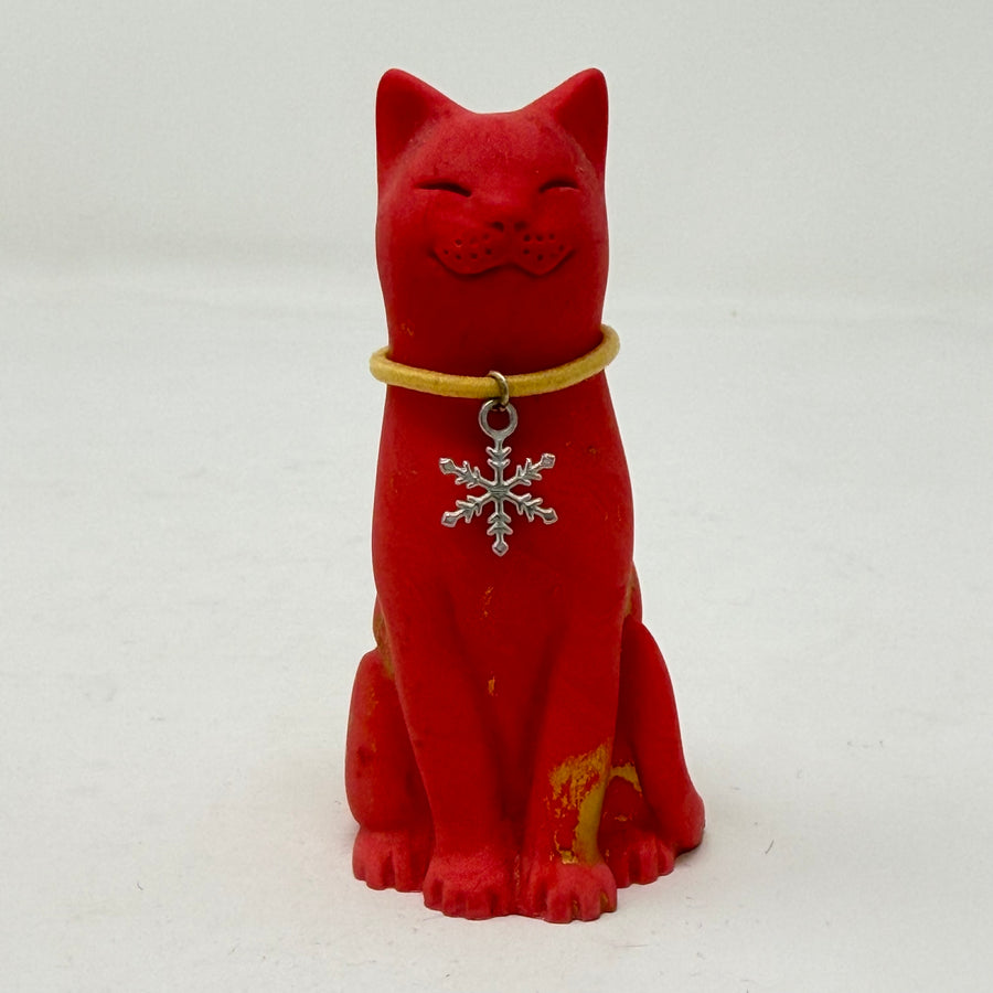 A red lucky cat figurine wearing a yellow collar with a silver snowflake pendant, set against a plain white background.