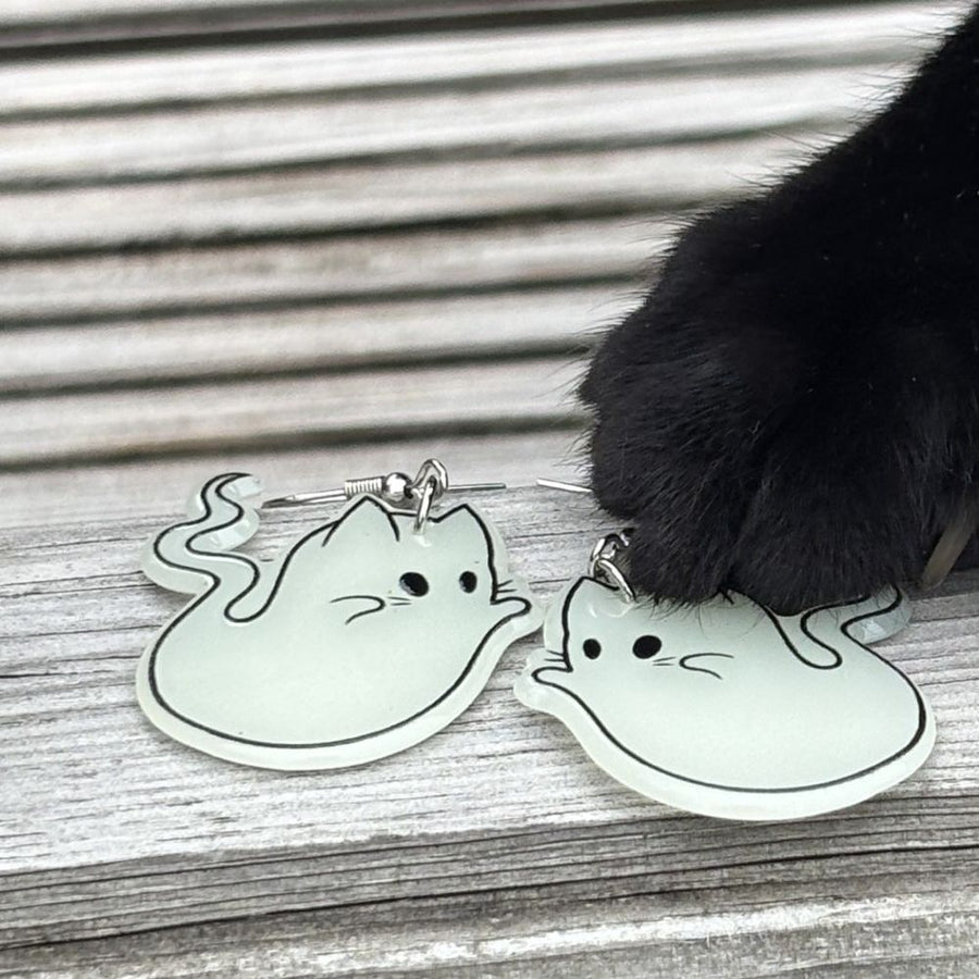 Glowing ghost-shaped earrings on a wooden surface with a cat's paw interacting with them