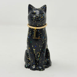 A black lucky cat figurine with gold holographic speckles, wearing a gold collar with a hanging letter 'L' charm