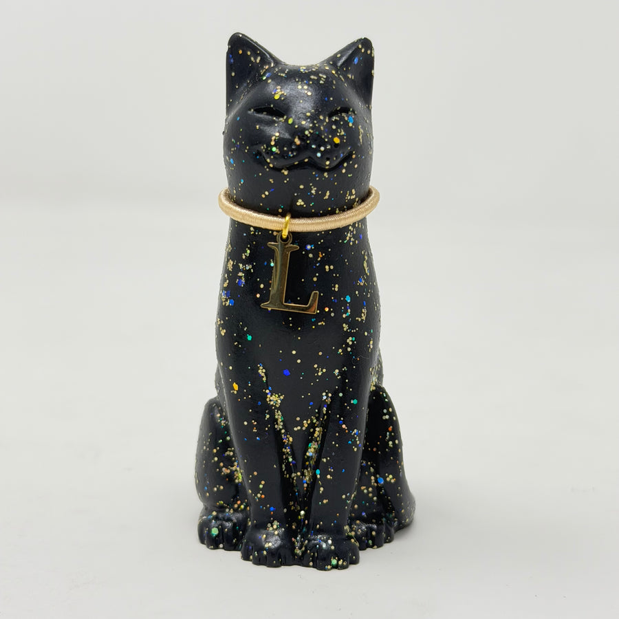 A black lucky cat figurine with gold holographic speckles, wearing a gold collar with a hanging letter 'L' charm