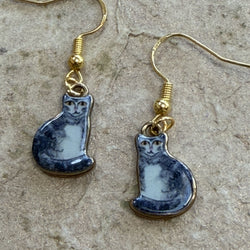 A pair of dangling earrings with gold-tone fishhook wires and small ceramic cat pendants in a grey-and-white painted style