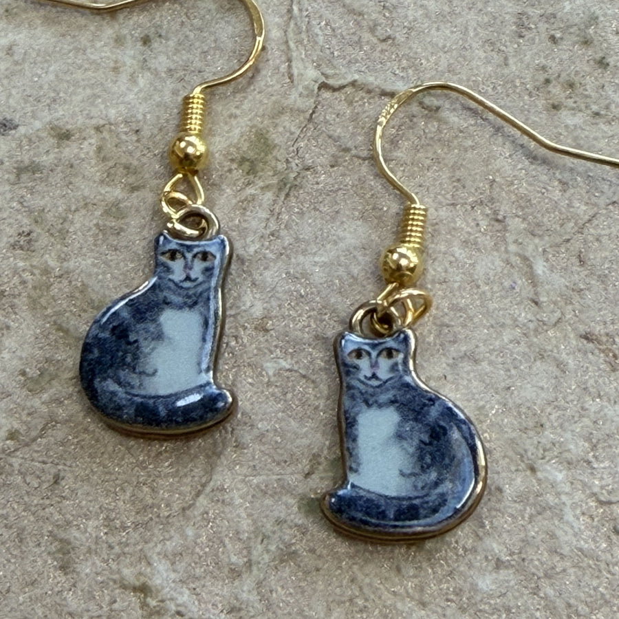 A pair of dangling earrings with gold-tone fishhook wires and small ceramic cat pendants in a grey-and-white painted style