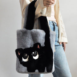 A person holds a grey fuzzy tote bag shaped like a black cat with large white eyes