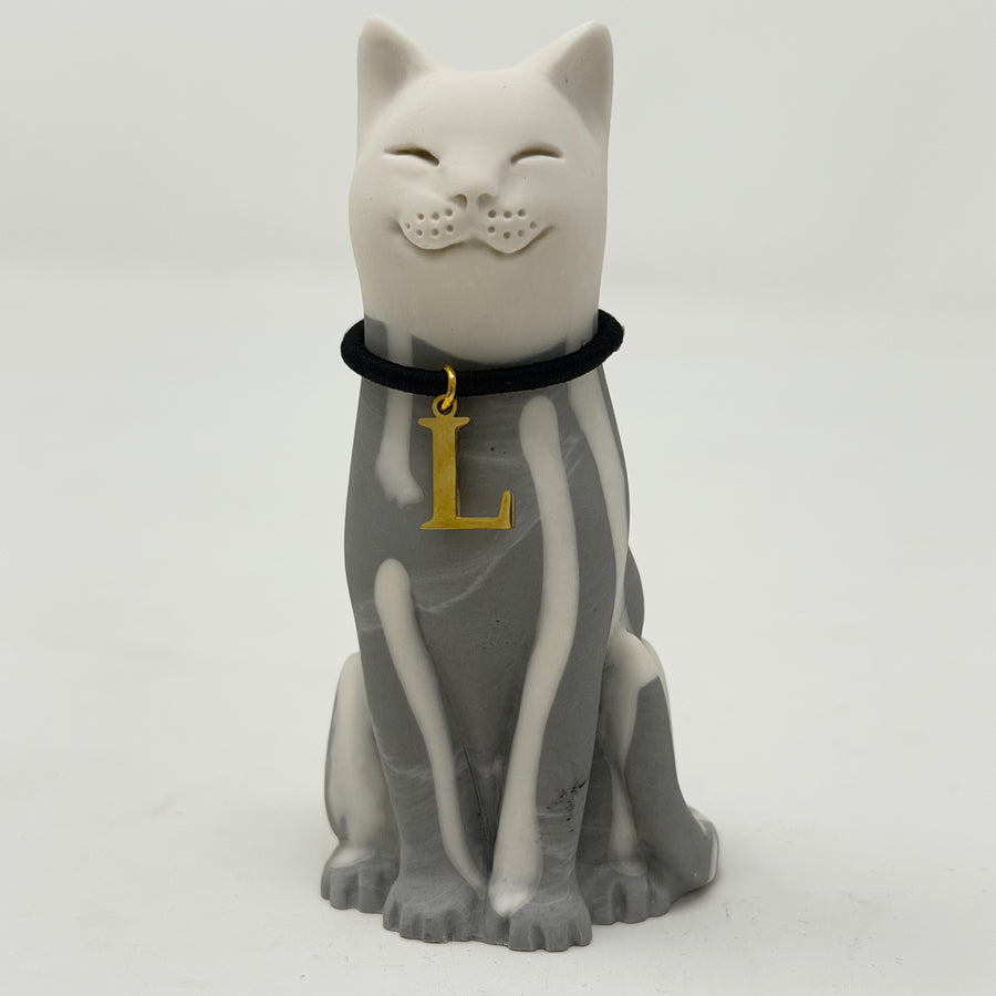 A resin lucky cat figurine in a sitting position, smiling, and featuring a grey and white pattern, black collar, and a gold letter 'L' charm