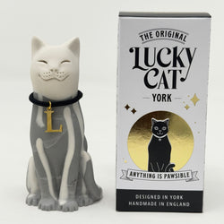 A white and grey lucky cat resin figurine wearing a black collar with a gold letter 'L' pendant, shown next to its retail packaging box, branded 'The Original Lucky Cat York'