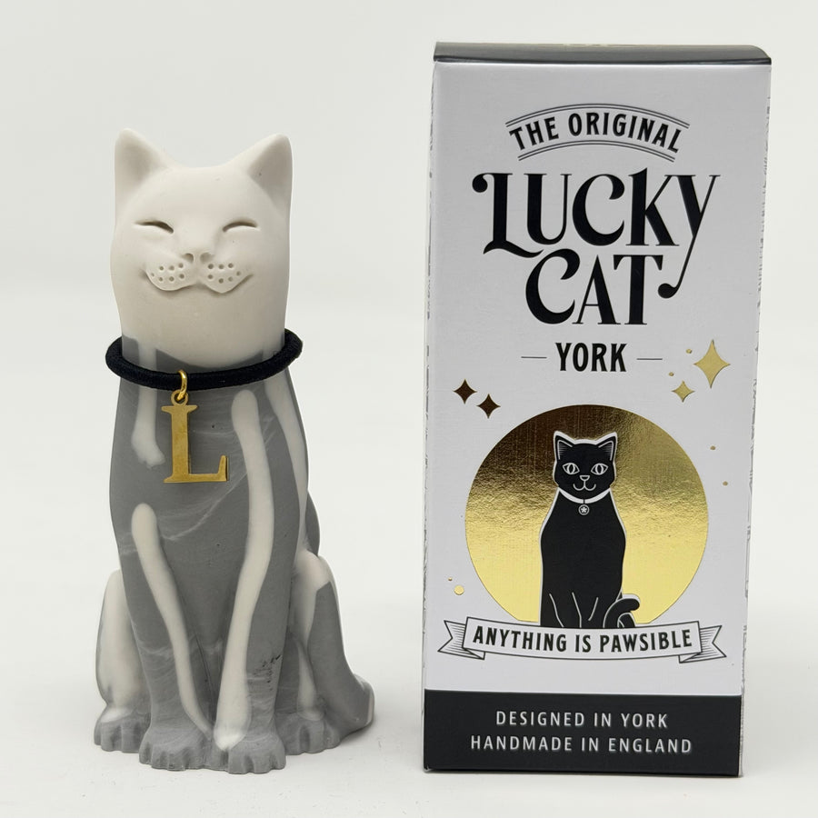 A white and grey lucky cat resin figurine wearing a black collar with a gold letter 'L' pendant, shown next to its retail packaging box, branded 'The Original Lucky Cat York'