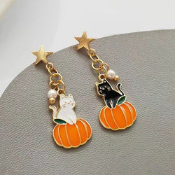 A pair of gold-tone dangle earrings with charms shaped like a white cat on an orange pumpkin and a black cat on a pumpkin