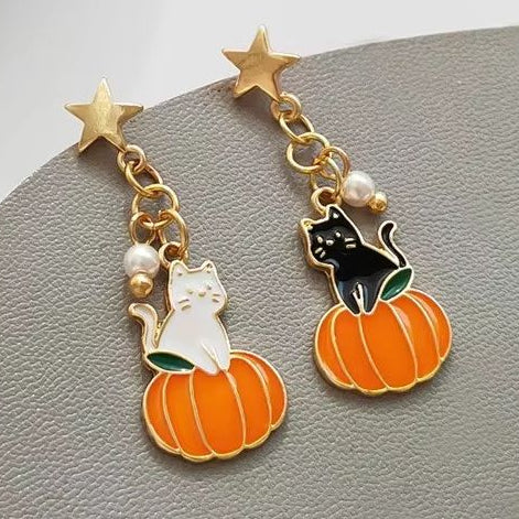 A pair of gold-tone earrings featuring star-shaped studs and charms of a white cat and a black cat on orange pumpkins