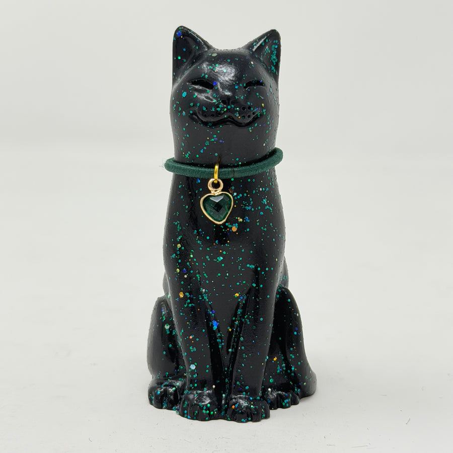 A glossy black lucky cat figurine with blue and green holographic speckles, wearing a dark green collar with a gold heart-shaped charm