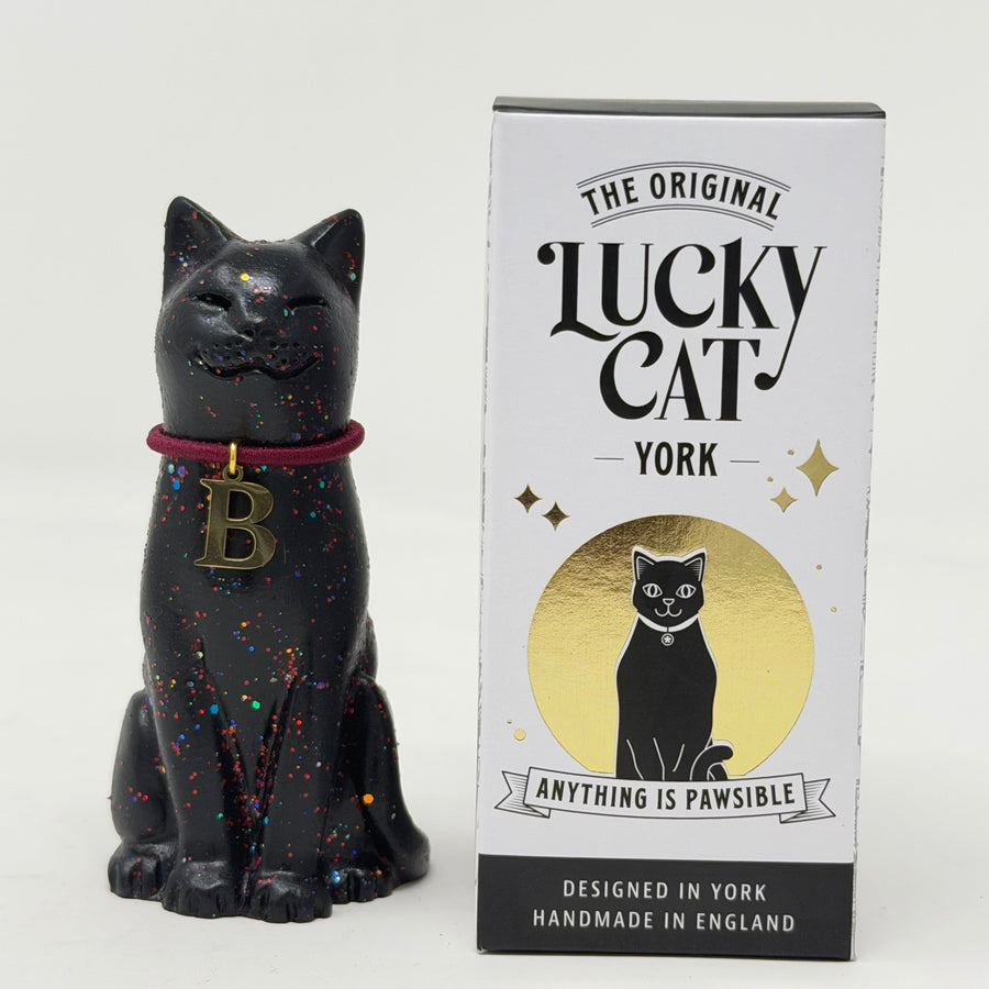 A black lucky cat figurine with colorful glitter speckles, a burgundy collar with a gold letter 'B', sitting next to its branded box labeled 'The Original Lucky Cat York'.
