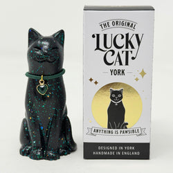 A black glittery lucky cat figurine wearing a green collar with a heart-shaped charm, displayed next to its white and gold branded box, 'The Original Lucky Cat York'.