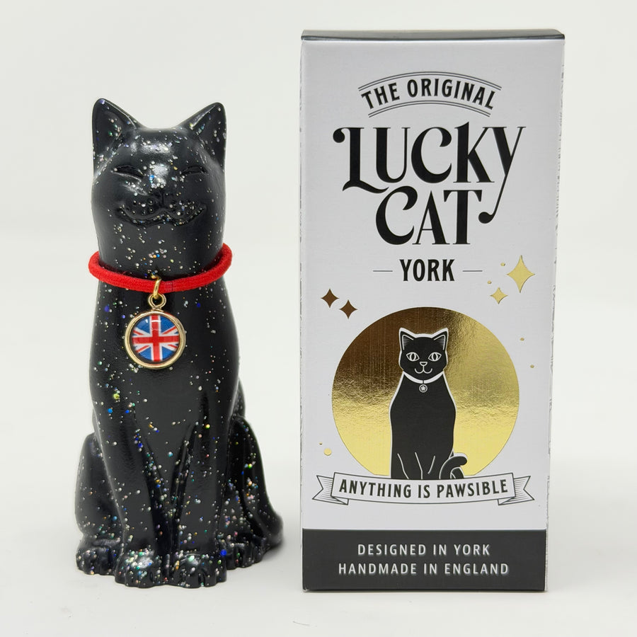 A black glittery lucky cat figurine wearing a red collar with a Union Jack charm, shown next to its white and gold retail box labeled 'The Original Lucky Cat York'.