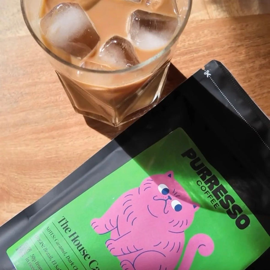 A black stand-up pouch of Purresso Coffee labelled 'The House Cat' next to a glass of iced coffee with ice cubes.