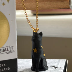 A black cat pendant necklace with gold star accents hanging from a gold chain, displayed next to its product box on a tabletop.