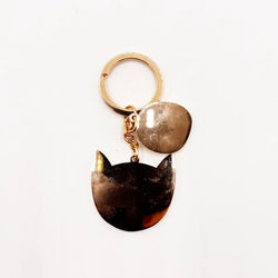 A gold-tone metal keychain featuring two dangling charms: one irregularly shaped metallic charm and one cat-head silhouette charm