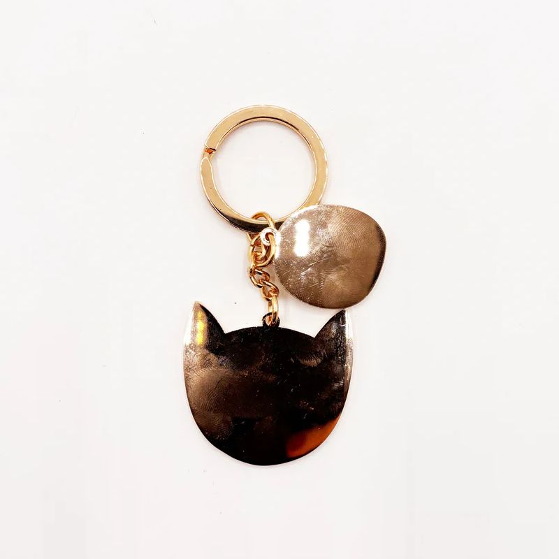A gold-tone metal keychain featuring two dangling charms: one irregularly shaped metallic charm and one cat-head silhouette charm