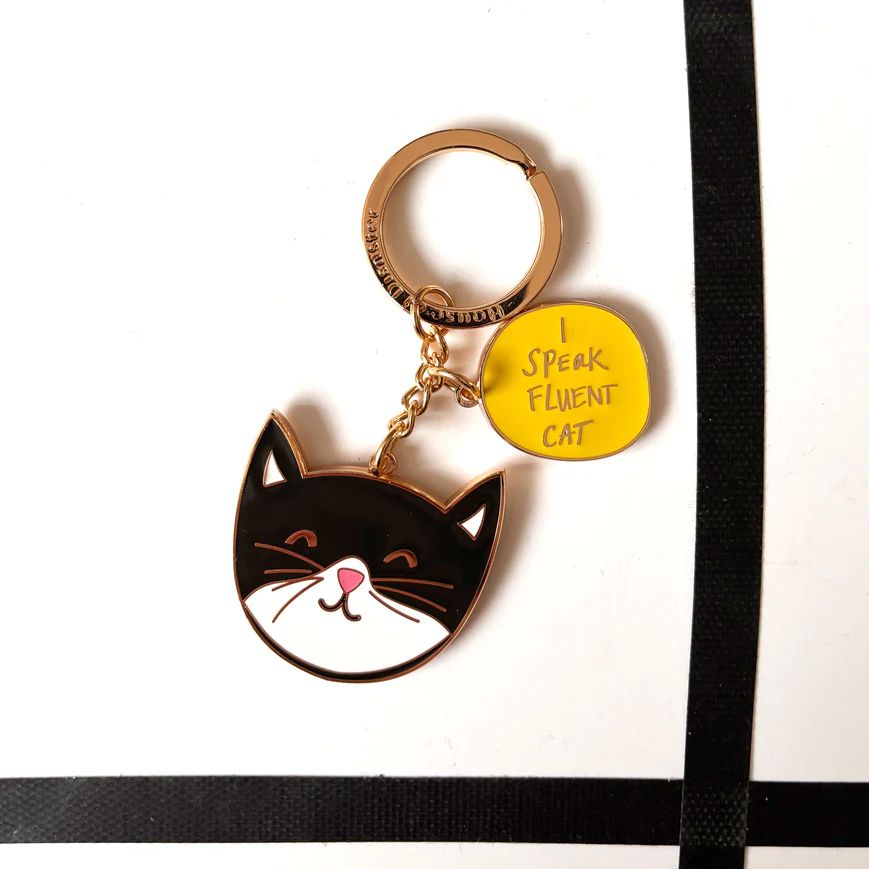 A black and white tuxedo cat enamel keychain featuring a small yellow charm that reads 'I SPEAK FLUENT CAT'