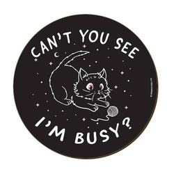 A round black coaster with a white line-art illustration of a cat playing with a ball of yarn, surrounded by the text 'CAN'T YOU SEE I'M BUSY?'