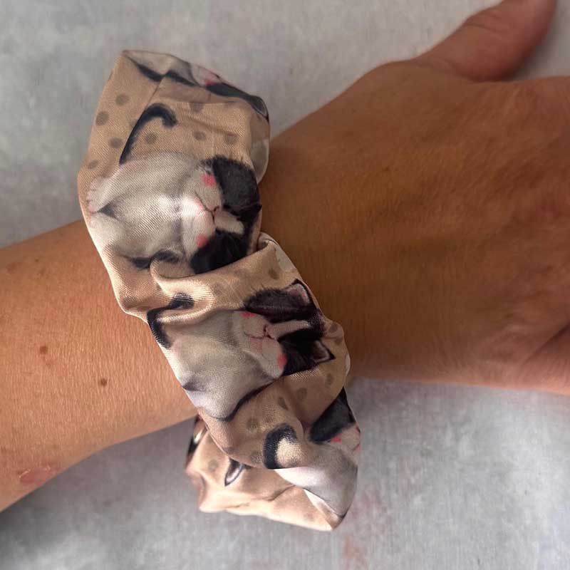 A fabric hair scrunchie with a printed pattern of sleeping cats, worn on a person's wrist against a light grey background.
