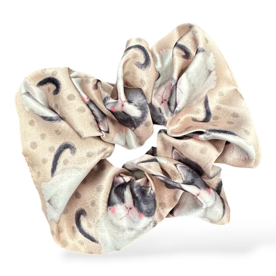 A fabric scrunchie with a printed pattern of sleeping cats in beige, grey, and white, set against a plain white background.