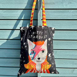 A black canvas tote bag featuring a stylized cat illustration with lightning-bolt makeup and the text 'KITTY STARDUST'