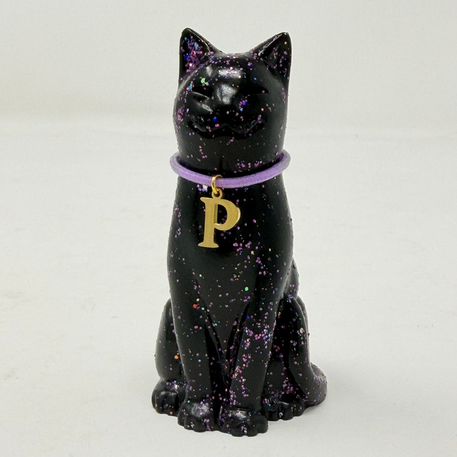 A black lucky cat figurine with purple holographic speckles, wearing a purple collar with a gold letter 'P' charm