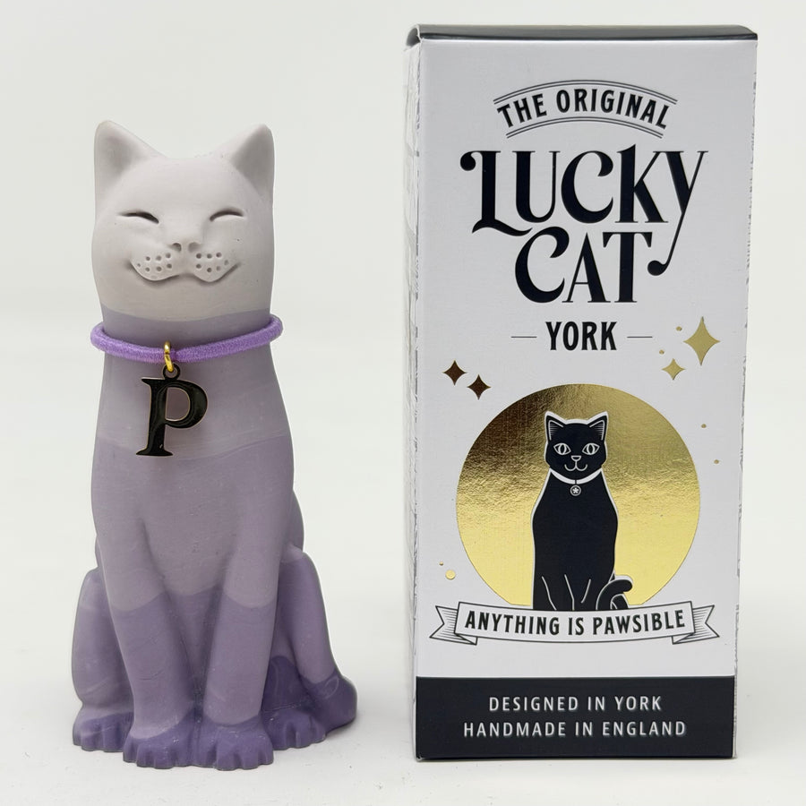 A lavender and white ombre lucky cat figurine wearing a purple collar with a black letter 'P' charm, standing by its branded box 'The Original Lucky Cat York'.