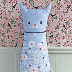 A light blue cat-shaped plush cushion with a floral pattern, featuring embroidered black eyes, white whiskers, and a small red nose, set against a pink floral wallpaper background.