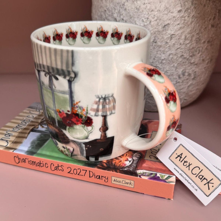 China mug with printed design of two tabby cats by a window. Mug is on top of an Alex Clark diary. Mug has an Alex Clark tag