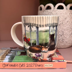 China mug with printed design of two tabby cats by a window. Mug is on top of an Alex Clark diary