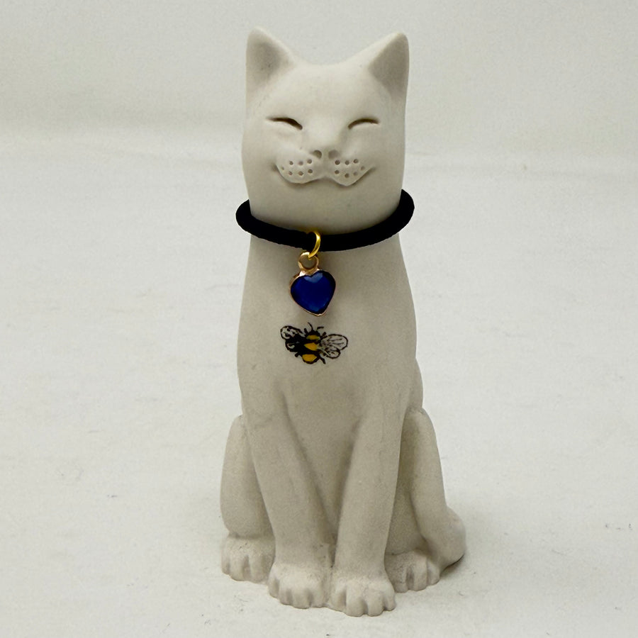 A white resin lucky cat figurine wearing a black collar with a blue heart-shaped pendant and a small bee decoration