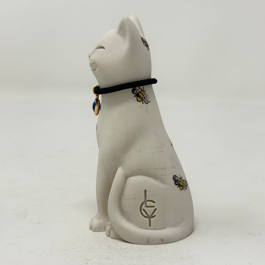 A white resin lucky cat figurine wearing a black collar with small blue and gold charm, decorated with tiny bee motifs