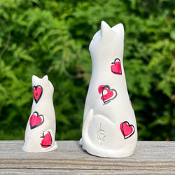 Two white resin cat figurines with hand applied red heart designs on a wooden surface with a green blurred background.