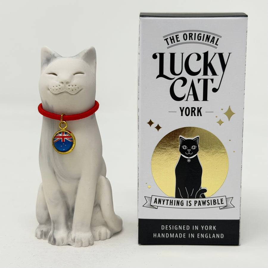 A greay and white resin Lucky Cat figurine wearing a red collar with a blue and gold flag charm, sitting next to its branded box "The Original Lucky Cat York"