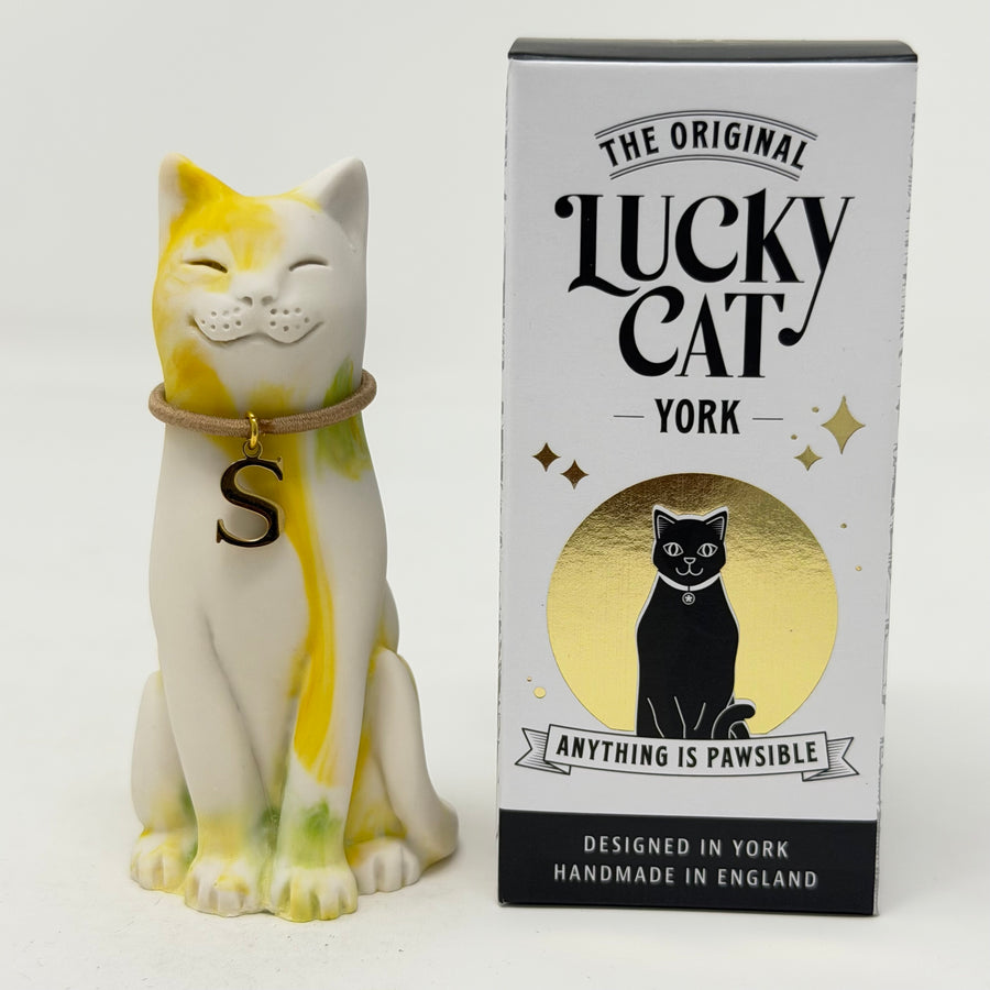 A resin Lucky Cat figurine with yellow and green marbled paint on a white base, wearing a brown collar with a gold 'S' charm, sitting next to its branded box "The Original Lucky Cat York"