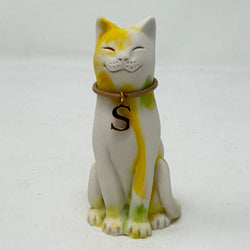 A resin Lucky Cat figurine with a white base and yellow and green paint details, wearing a brown collar with a black letter 'S' charm, with a smiling expression