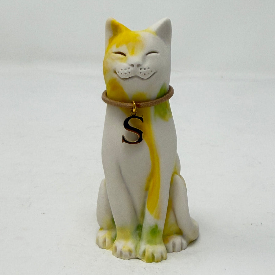 A resin Lucky Cat figurine with a white base and yellow and green paint details, wearing a brown collar with a black letter 'S' charm, with a smiling expression