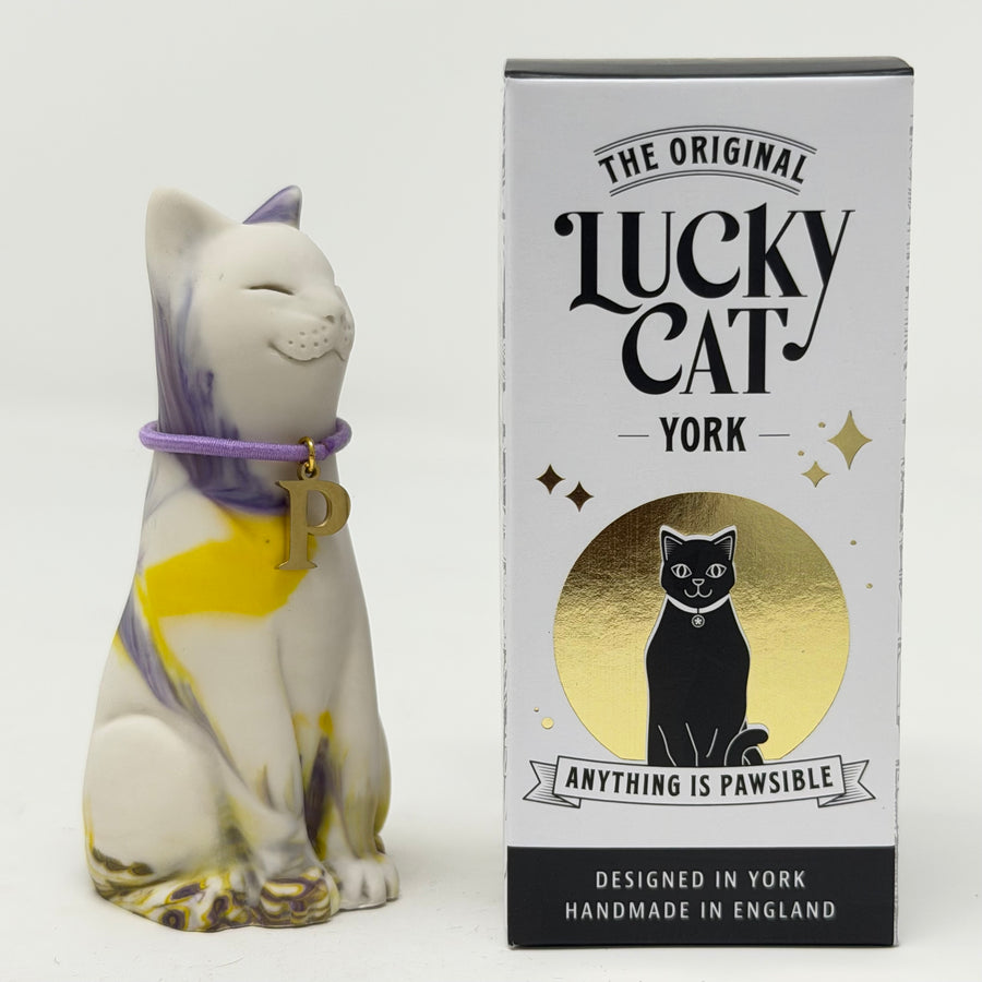 A ceramic Lucky Cat figurine with a white body, purple and yellow stripes, and a purple collar with a gold letter 'P' charm, shown next to its white and gold branded packaging box.